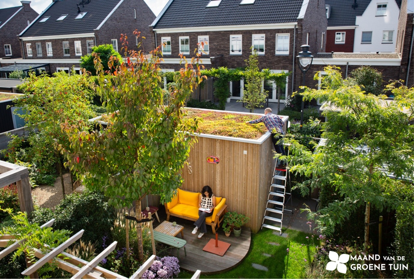 Foto van een groene tuin, met schuur met groen dak. Een man staat op een ladder om het groene dak te onderhouden, terwijl een vrouw een boek leest op een gele bank.